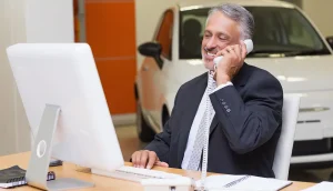 Mature insurance agent in a suit smiling while talking on the phone with a customer at his desk in front of a computer