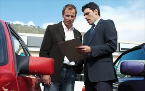 Car dealer in suit showing clipboard to client during used car appraisal outside dealership