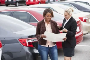 Woman reading vehicle documents with a dealership representative in a car lot surrounded by multiple parked cars