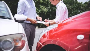 Two men standing between a red and white car, filling out an accident claim form after a minor crash