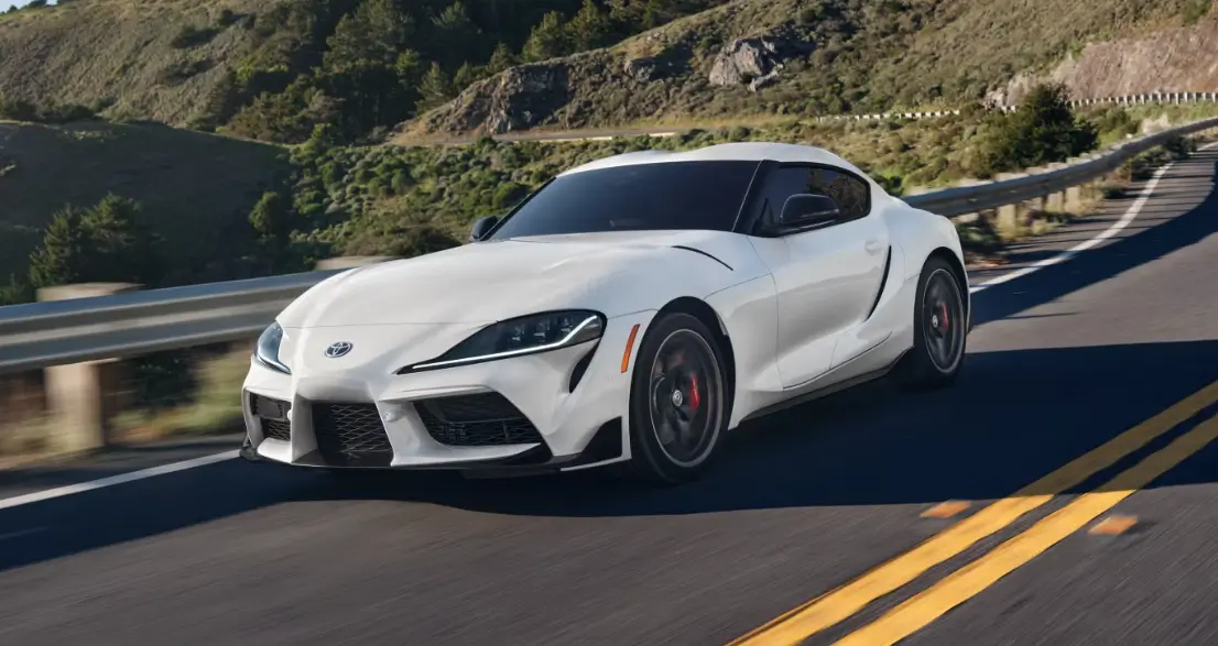 White Toyota Supra sports car speeding on a scenic mountain road, highlighting its sleek design and performance.