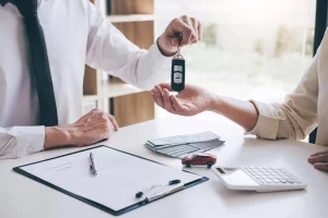 Car buyer receiving keys at dealership with money, paperwork, and calculator on table.