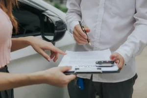 Close-up of two people reviewing and signing a document on a clipboard next to a white car.