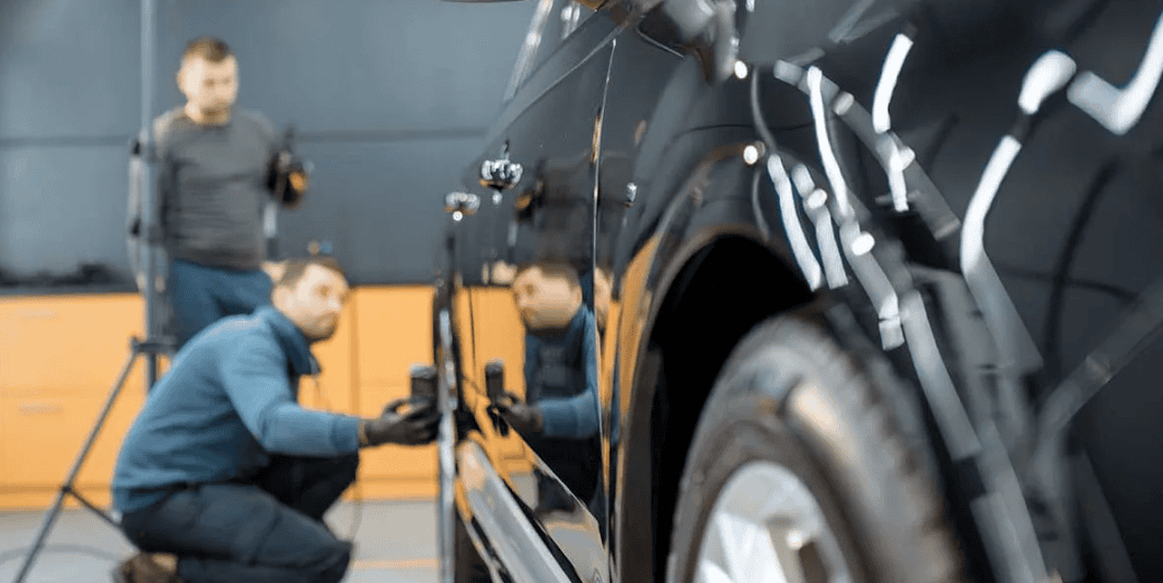 Two auto body technicians using inspection lights to examine a dark vehicle's body panel finish and repair quality in a professional shop