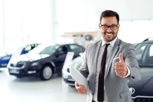 Confident car salesman in a gray suit smiling and giving a thumbs up inside a bright vehicle showroom