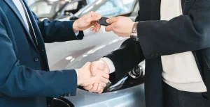 Two men shaking hands and exchanging car keys in front of a silver vehicle at a dealership