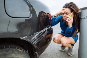 Woman on phone looking at deep scratch on her black car, appearing distressed after a parking accident