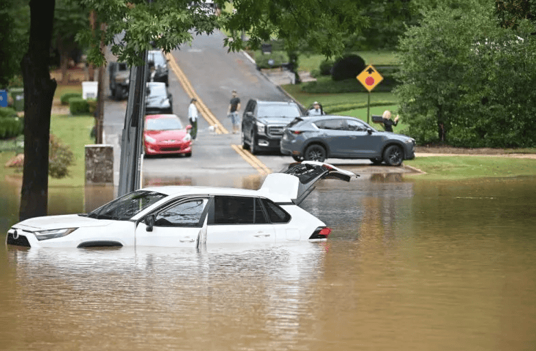White car submerged in floodwater on a residential street with bystanders and other vehicles visible in the background.