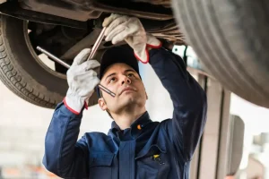 Focused auto mechanic working underneath a lifted car, performing an undercarriage inspection with metal tools