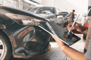 Insurance adjuster using a tablet to assess front-end collision damage on a car at a repair shop, estimating total loss or repair costs
