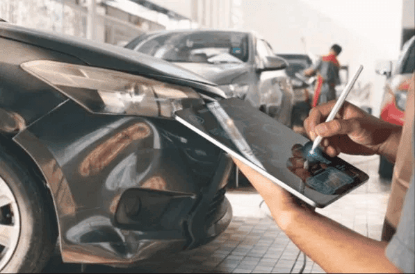 Insurance adjuster using a tablet to assess front-end collision damage on a car at a repair shop, estimating total loss or repair costs