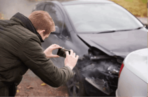 Man photographing front-end collision damage with a smartphone, capturing insurance claim photo evidence at the scene