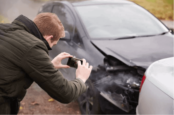 Man photographing front-end collision damage with a smartphone, capturing insurance claim photo evidence at the scene