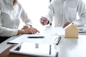 Two people reviewing insurance documents and signing claim forms on a desk with a small house model and tablet