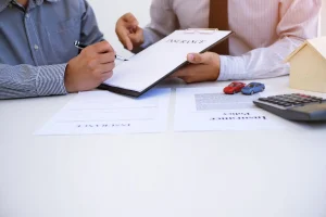 Two people signing car insurance documents at a desk with model cars, calculator, and miniature house