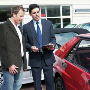 Car dealer in suit showing clipboard to male client next to a red car during vehicle appraisal