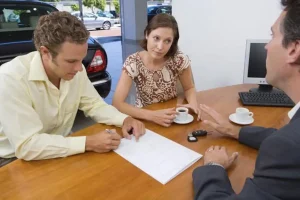 Couple discussing paperwork with a representative at a dealership or insurance office, signing documents next to car keys and coffee cups