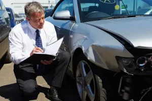 Insurance adjuster kneeling to inspect front-side collision damage on a silver vehicle